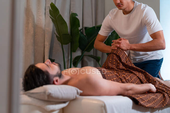 Vista lateral del masajista de cultivos anónimo haciendo masaje tailandés para el cliente masculino en el moderno salón de spa - foto de stock