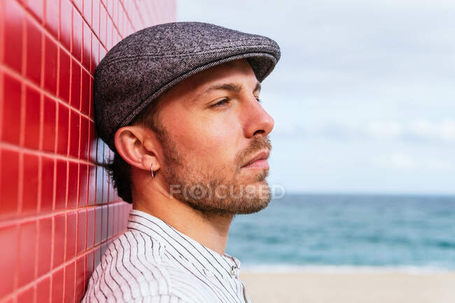 Side view of thoughtful young bearded male in stylish striped shirt and hat standing looking away near red wall and enjoying sunny summer day on street — Stock Photo