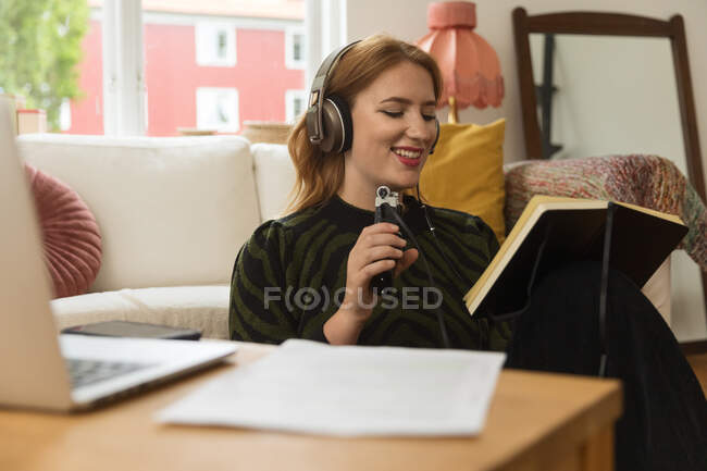 Восхитительная женщина-радиоведущий записи подкаста при использовании микрофона и чтения заметки из бумаги дома — стоковое фото