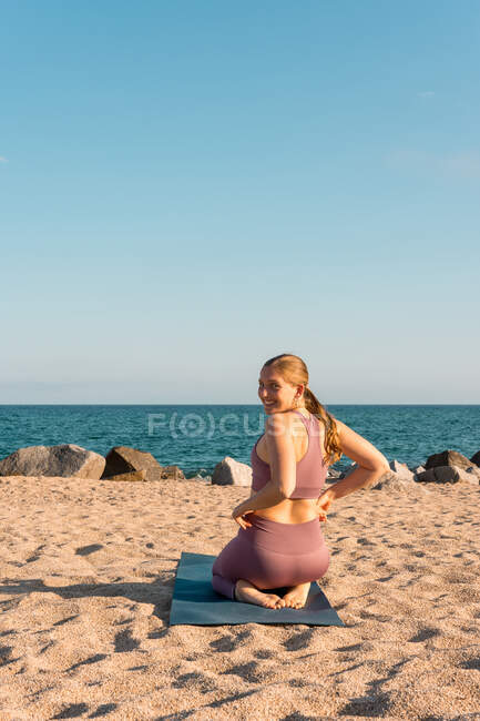 Vue arrière de la femme souriante assise sur le tapis tout en se préparant à pratiquer le yoga sur le bord de la mer et en regardant la caméra — Photo de stock