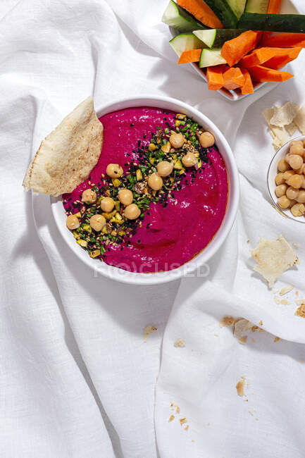 Top view of appetizing beetroot hummus garnished with chickpea served on fabric background with bread and fresh carrot and cucumber sticks — Stock Photo