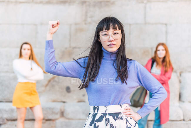 Confident ethnic Asian woman showing bicep while standing against group of multiracial females showing concept of girl power — Stock Photo