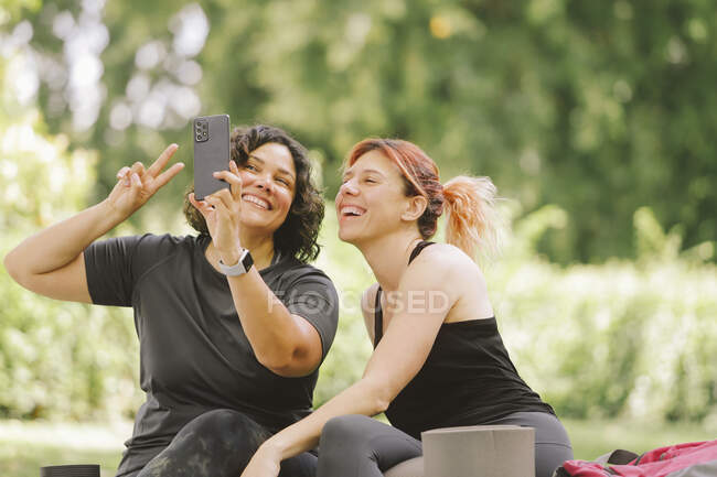 Joyeux jeunes amies multiraciales en vêtements décontractés souriant et montrant geste de victoire tout en prenant selfie sur smartphone reposant dans un jardin vert le jour ensoleillé — Photo de stock