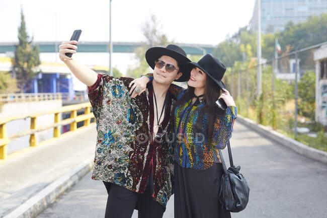 Coppia positiva innamorata in elegante abbigliamento hipster abbracciando in strada e prendendo auto colpo sul telefono cellulare — Foto stock