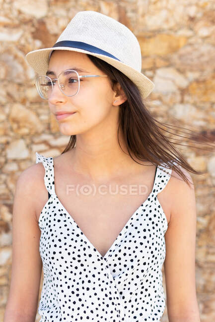Молодая серьезная женщина в шляпе и очках смотрит в сторону каменной стены в летний день — стоковое фото