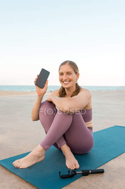 Delighted female in activewear sitting on yoga mat demonstrating mobile phone on seashore — Stock Photo