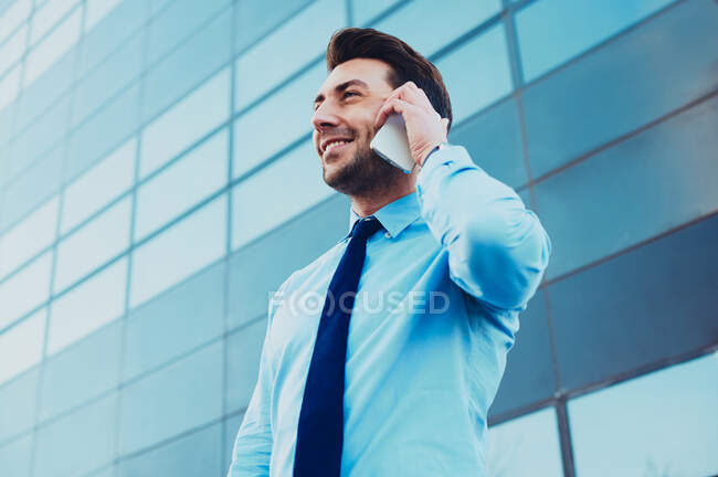 Знизу стильний щасливий керівник чоловічої статі в офіційному одязі, що розмовляє на мобільному телефоні, дивлячись далеко в місто — стокове фото