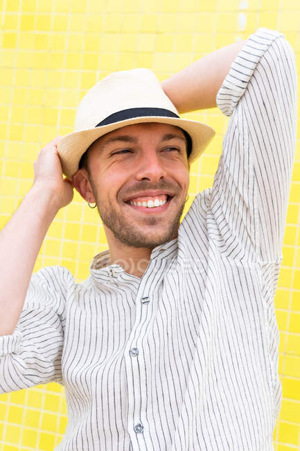 Feliz jovem barbudo hipster cara na roupa da moda e chapéu mantendo as mãos atrás da cabeça e sorrindo enquanto refrigerando sozinho contra a parede amarela no dia de verão — Fotografia de Stock