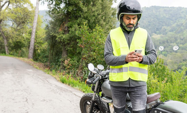 Thoughtful adult Hispanic male biker in protective helmet and vest messaging on mobile phone while standing near broken motorbike near lush green woods — Stock Photo