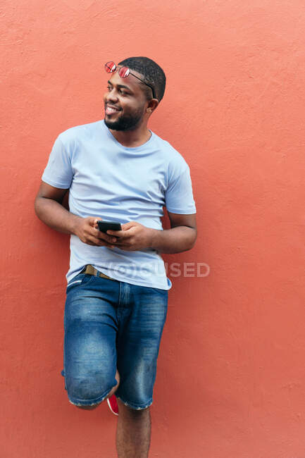 Uomo africano sorridente con uno smartphone che distoglie lo sguardo mentre si trova in città — Foto stock