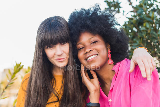Lächelndes multirassisches Paar lesbischer Frauen, die sich umarmen, während sie zusammen auf der Straße sitzen und in die Kamera schauen — Stockfoto