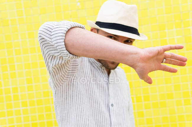 Unrecognizable guy in stylish summer outfit and hat doing no gesture and hiding face from camera against yellow wall — Stock Photo