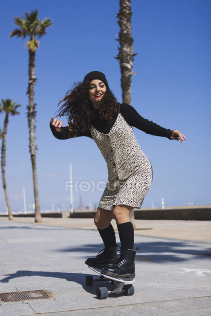 Все тело активной счастливой женщины в платье на скейтборде по дороге вдоль песчаного пляжа и высокие ладони во время тренировки — стоковое фото