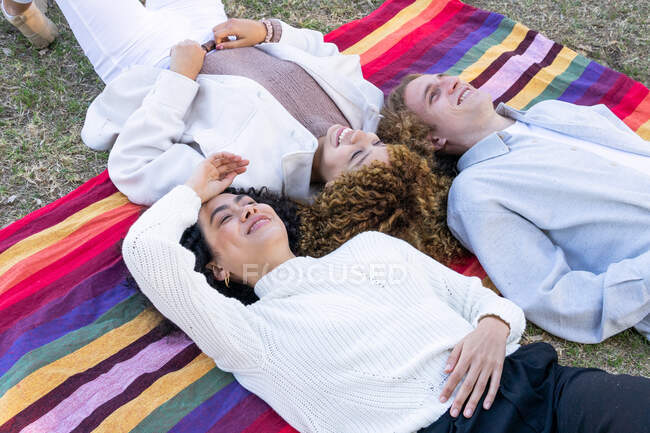 High angle of diverse women and man with curly hair lying face to face on colorful plaid in park looking up — Stock Photo