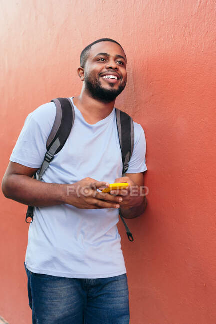 Homem com mochila e telefone celular olhando para longe enquanto se inclina na parede — Fotografia de Stock