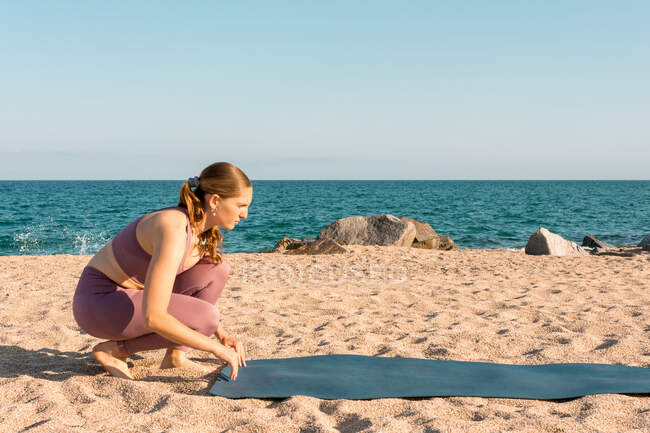 Full body side view of young female in sportswear placing yoga mat on sand while preparing for practice on beach near ocean — Stock Photo