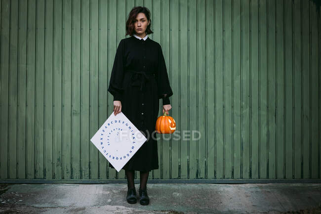 Femme sérieuse en robe noire debout avec citrouille illuminée et planche d'esprit contre un mur en bois et regardant la caméra pendant la célébration d'Halloween — Photo de stock