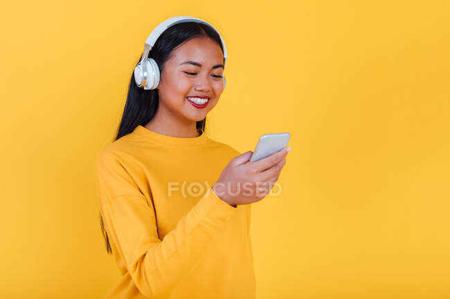 Content Asian female listening to music in headphones and taking self shot on smartphone on yellow background in studio — Stock Photo