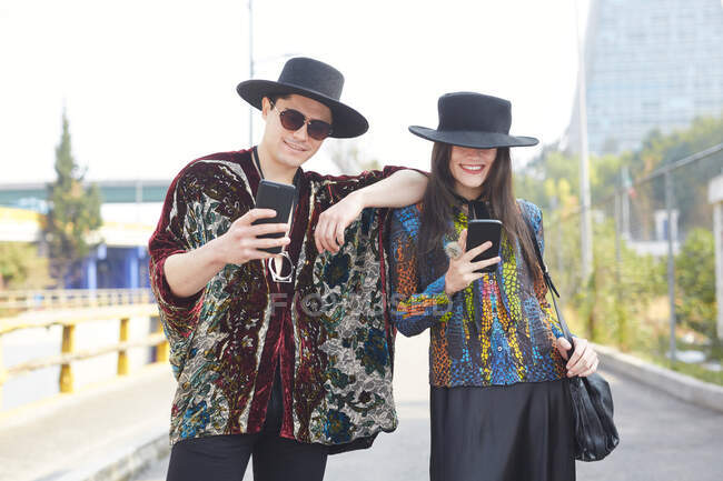Couple enchanté en vêtements tendance hipster debout en ville et utilisant des téléphones mobiles en été — Photo de stock