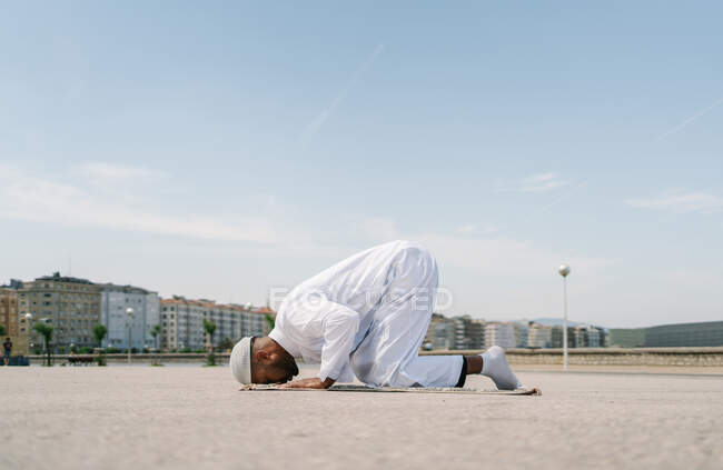 Мусульманский мужчина стоит на коленях на ковре и касается земли лбом во время молитвы на песчаном пляже в солнечный день — стоковое фото