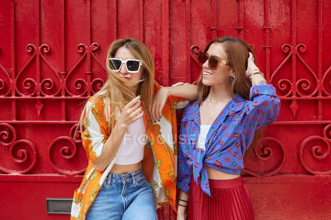 Amigos alegres do sexo feminino em roupas da moda e óculos de sol em pé contra a porta vermelha na cidade no verão — Fotografia de Stock