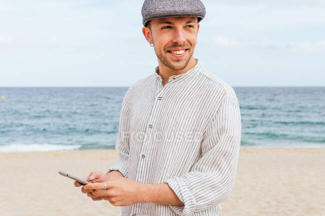 Joven barbudo positivo con camisa elegante y gorra sonriendo y mirando hacia otro lado mientras navega por el teléfono móvil en la playa de arena cerca del mar - foto de stock