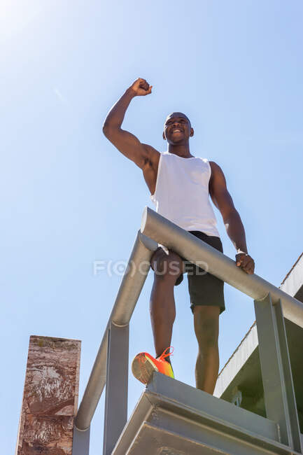 D'en bas de l'athlète masculin afro-américain en forme debout sur la terrasse avec les premières et célébrer la victoire tout en profitant du triomphe — Photo de stock