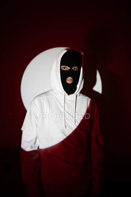Unrecognizable serious rebellious young male in black balaclava mask and white hoodie standing near wall in sunlight — Stock Photo
