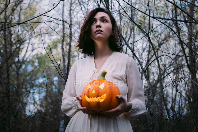 D'en bas femelle en robe blanche debout avec lanterne de citrouille rayonnante dans les bois sur Halloween et détournant les yeux — Photo de stock