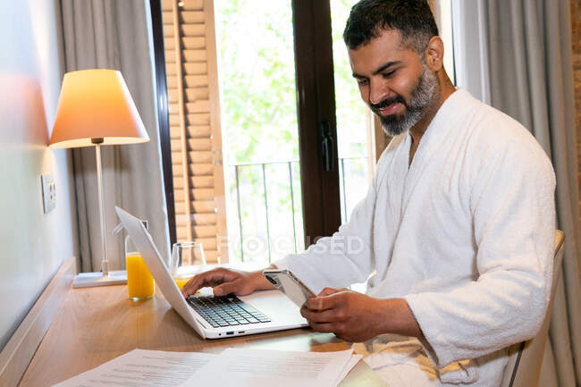 Vista laterale positivo imprenditore etnico maschile seduto a tavola con telefono cellulare e laptop collocato vicino a documenti e succo di frutta — Foto stock
