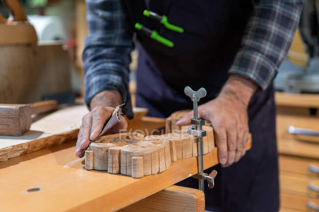 Ernte anonyme männliche Holzarbeiter mit Metallklammer geschnitzte Holzdetails auf Werkbank zu fixieren, während Feilen mit Schleifpapier in Tischlerei Werkstatt — Stockfoto