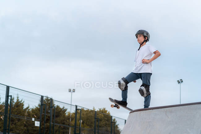Patineur adolescent en équipement de protection équitation skateboard pendant le week-end dans le skate park et en regardant ailleurs — Photo de stock