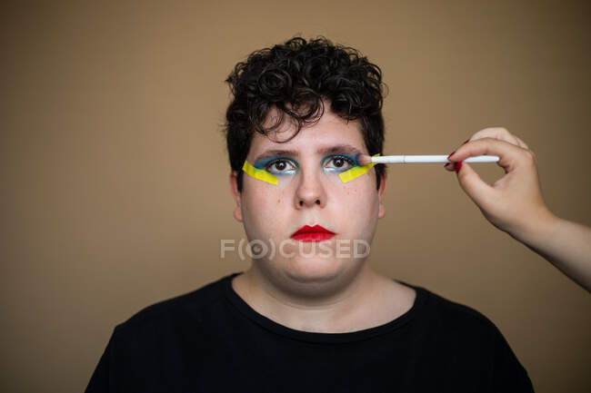 Estilista de colheita com escova aplicando maquiagem brilhante nas pálpebras de macho queer olhando para a câmera — Fotografia de Stock