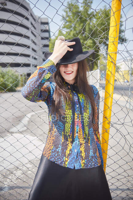 Conteúdo fêmea em roupas da moda em pé perto de cerca de malha na cidade no dia ensolarado e cobrindo os olhos com chapéu — Fotografia de Stock