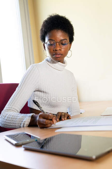 Black female physician writing information on paper sheet while preparing medical report at table in office of modern clinic — Stock Photo