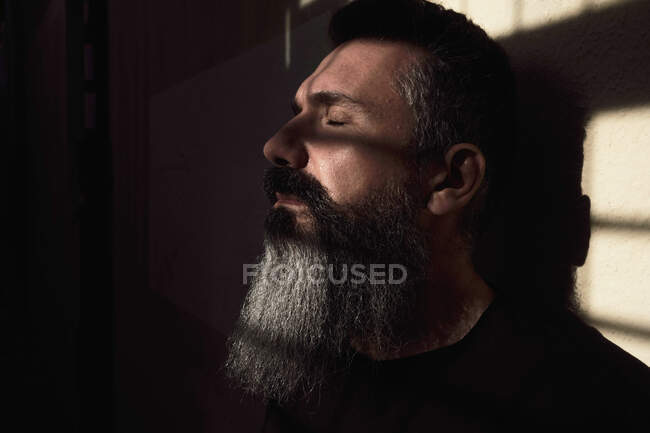 Вид збоку мрійливого зрілого бородатого чоловіка з закритими очима в темній кімнаті з сонячним світлом — стокове фото