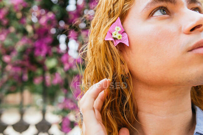 Dreamy curious male with long wavy hair standing looking away in street in summer in park under blossoming tree with pink flowers — Stock Photo