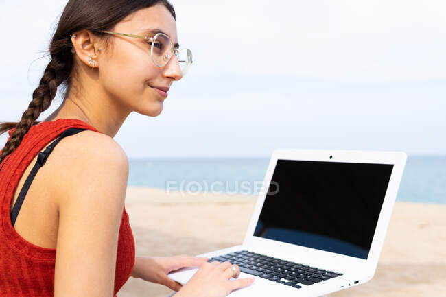 Содержание женщины-фрилансера, сидящей на берегу моря и печатающей на ноутбуке, работая удаленно над проектом летом — стоковое фото