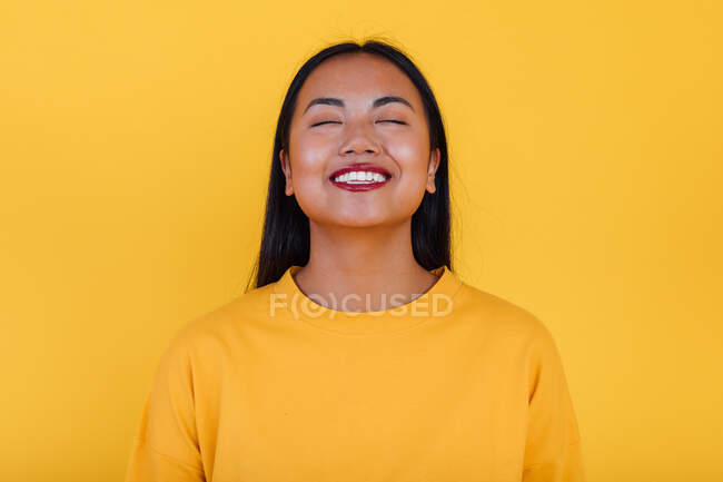 Encantado Asiático fêmea em pé no fundo amarelo no estúdio com os olhos fechados — Fotografia de Stock