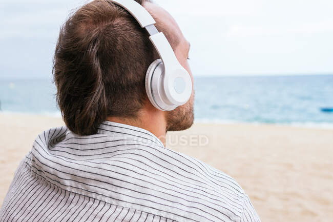 Back view of unrecognizable carefree young bearded guy in stylish casual shirt listening to music through wireless headphones and enjoying fresh breeze while spending summer day on sandy beach near sea — Stock Photo