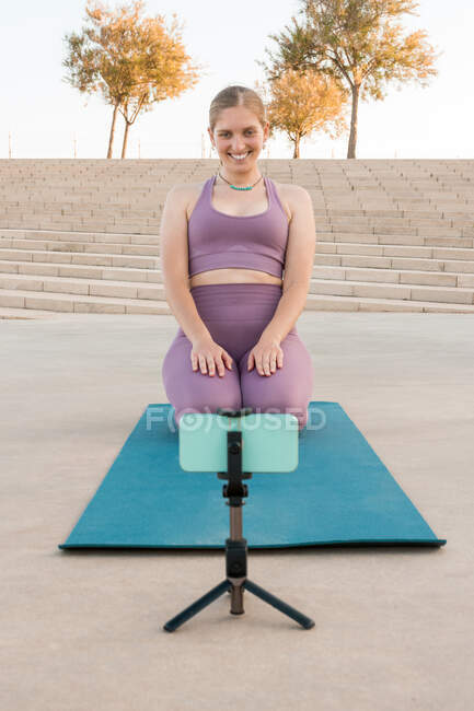 Mujer alegre sentada en la esterilla practicando yoga con teléfono móvil - foto de stock