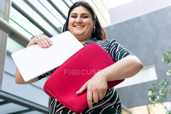 From below of positive plus size young female student in stylish outfit taking laptop of red case on urban background — Stock Photo