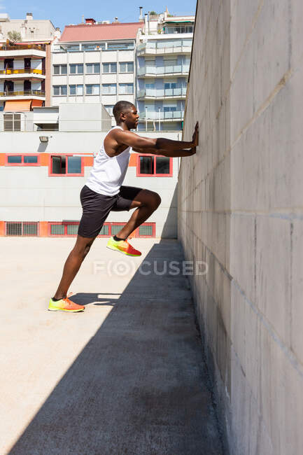 Vista laterale dell'atletico maschio afroamericano appoggiato alla parete e che corre sul posto durante l'allenamento attivo nella giornata di sole sul campo sportivo — Foto stock