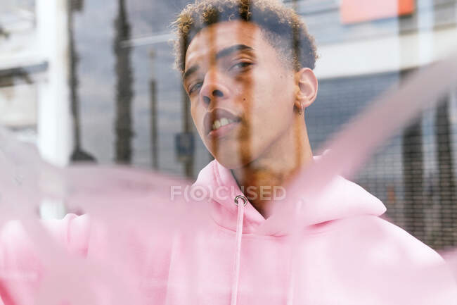 Grâce à un verre de jeune homme hipster ethnique sérieux avec une coiffure afro vêtue d'un sweat à capuche rose à la fenêtre et regardant la caméra — Photo de stock