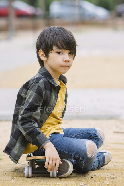Vista laterale corpo pieno di etnico ispanico ragazzo in abiti casual guardando la fotocamera mentre seduto su skateboard per strada — Foto stock
