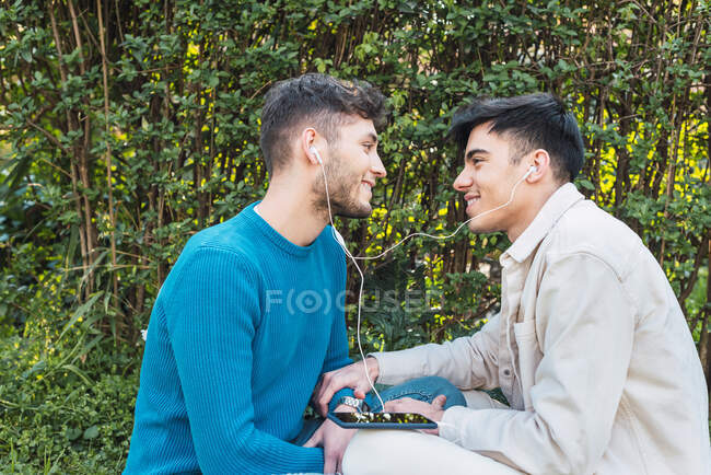 Вид сбоку улыбающейся пары гомосексуалистов, сидящих на лавочке в парке и наслаждающихся солнечным днем, слушая музыку и глядя друг на друга — стоковое фото