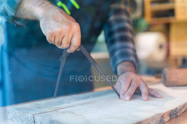 Crop unrecognizable male woodworker using professional compass or divider while marking wooden plank at workbench in carpentry workshop — Stock Photo