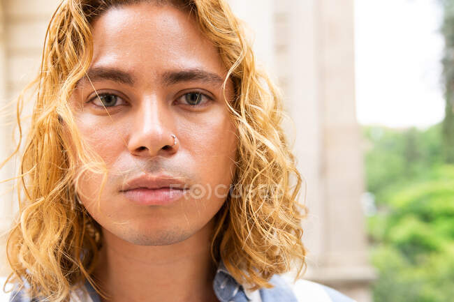Confident male in stylish summer clothes and with long wavy hair looking at camera in street — Stock Photo