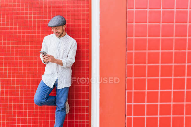Зміст молодого бородатого хіпстера в повсякденній сорочці поло і кепці перегляду мобільного телефону, стоячи на стіні червоного кольору на сонячному світлі — стокове фото