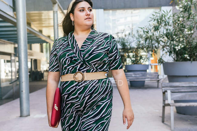 Современная молодая деловая леди в стильном платье с геометрической печатью и поясом с чехлом для ноутбука во время прогулки по городу — стоковое фото